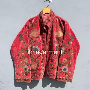 May include: A red velvet jacket with intricate floral embroidery in shades of gold, green, and blue. The jacket features a high collar and is displayed on a wooden hanger. The text "ilmagarment" is visible on the front.
