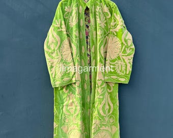 May include: A lime green velvet coat with intricate beige floral embroidery. The long coat is hanging on a wooden hanger against a dark blue background. The coat has long sleeves and a floral patterned lining.