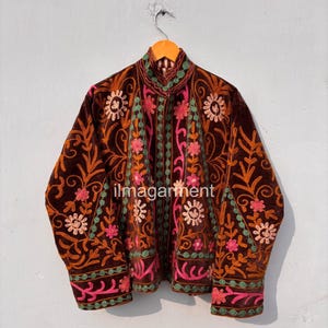 May include: A brown velvet jacket with intricate floral embroidery in shades of pink, orange, and green. The jacket features a high collar and long sleeves, with the text "ilmagarment" visible on the front.