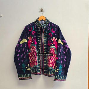 May include: A dark blue jacket featuring vibrant multi-coloured embroidery of two peacocks, floral motifs, and geometric patterns. The garment has a high collar, long sleeves, and a front opening. The text ilmagarment is visible on the front.