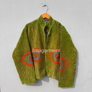 Könnte beinhalten: Eine grüne Samtjacke mit gestickten Augenmotiven in Rot, Blau und Braun. Die Jacke hat einen Stehkragen und lange Ärmel. Der Text "ilmagarment" ist auf der Vorderseite sichtbar.