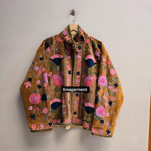 以下が含まれることがあります： ハイカラーと長袖が特徴のマスタードイエローのジャケット。ピンク、ダークブルー、ライトブルーの色合いの複雑な花柄刺繍が施されています。衣服の中央前面には「ilmagarment」という文字が黒でプリントされています。