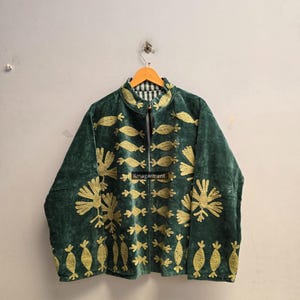 Op de afbeelding: Een donkergroene fluwelen jas met goudkleurige geborduurde patronen van gestileerde vogels en geometrische vormen. Het kledingstuk heeft een hoge kraag, lange mouwen en een opening aan de voorzijde. Een zwart label met de tekst ilmagarment is zichtbaar.