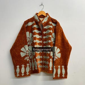 Puede incluir: Chaqueta de terciopelo color óxido con intrincados bordados florales verde claro. La chaqueta tiene cuello alto y mangas largas. El texto "llmagarment" es visible. La chaqueta está colgada de una percha de madera.