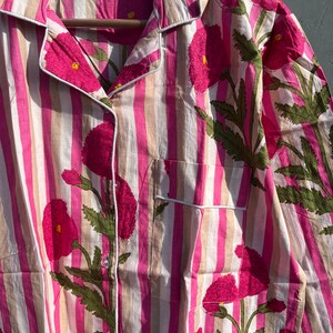 Puede incluir: Una camisa a rayas rosa y beige con un estampado floral de flores rosas y hojas verdes. La camisa tiene un cuello abotonado y un bolsillo en el lado izquierdo. La etiqueta de la camisa dice "100% algod&oacute;n Hecho en India" y "M".