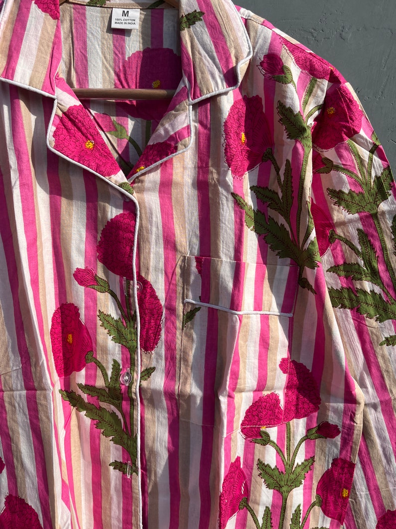 Puede incluir: Una camisa a rayas rosa y beige con un estampado floral de flores rosas y hojas verdes. La camisa tiene un cuello abotonado y un bolsillo en el lado izquierdo. La etiqueta de la camisa dice "100% algod&oacute;n Hecho en India" y "M".