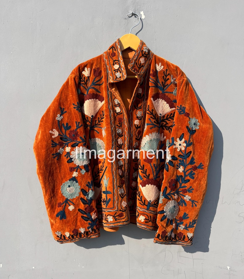 May include: A burnt orange velvet jacket with intricate floral embroidery in shades of blue, green, and pink. The jacket has a mandarin collar and a button closure.