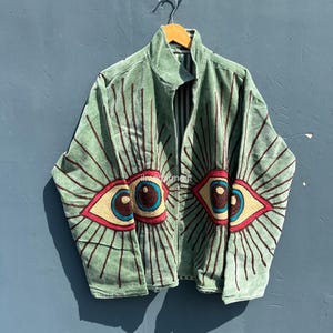 May include: A sage green zip-up jacket with embroidered eye designs. The eyes have blue irises, yellow and red accents, and brown radiating lines. The jacket is hanging on a wooden hanger against a blue background.