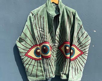 Könnte beinhalten: Eine salbeigrüne Jacke mit Reißverschluss und gestickten Augendesigns. Die Augen haben blaue Iris, gelbe und rote Akzente und braune Strahlenlinien. Die Jacke hängt an einem Holzbügel vor blauem Hintergrund.