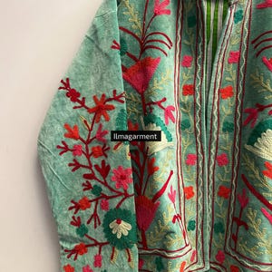 May include: A light green velvet jacket with intricate floral embroidery in shades of red, pink, and gold. The jacket features a mandarin collar and a striped green and white lining. The text "ilmagarment" is visible on the bottom of the jacket.