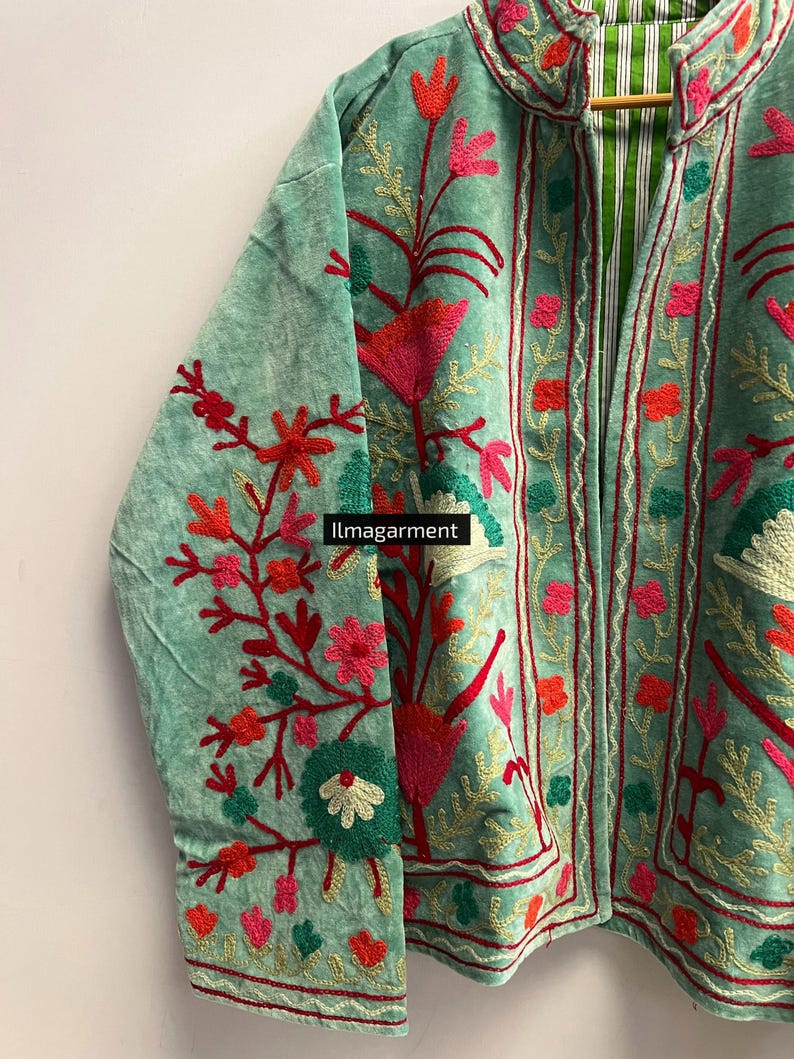 May include: A light green velvet jacket with intricate floral embroidery in shades of red, pink, and gold. The jacket features a mandarin collar and a striped green and white lining. The text "ilmagarment" is visible on the bottom of the jacket.