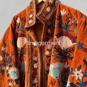 May include: A long, orange velvet jacket with intricate floral embroidery in shades of blue, pink, and brown. The jacket has a stand-up collar and a button closure.