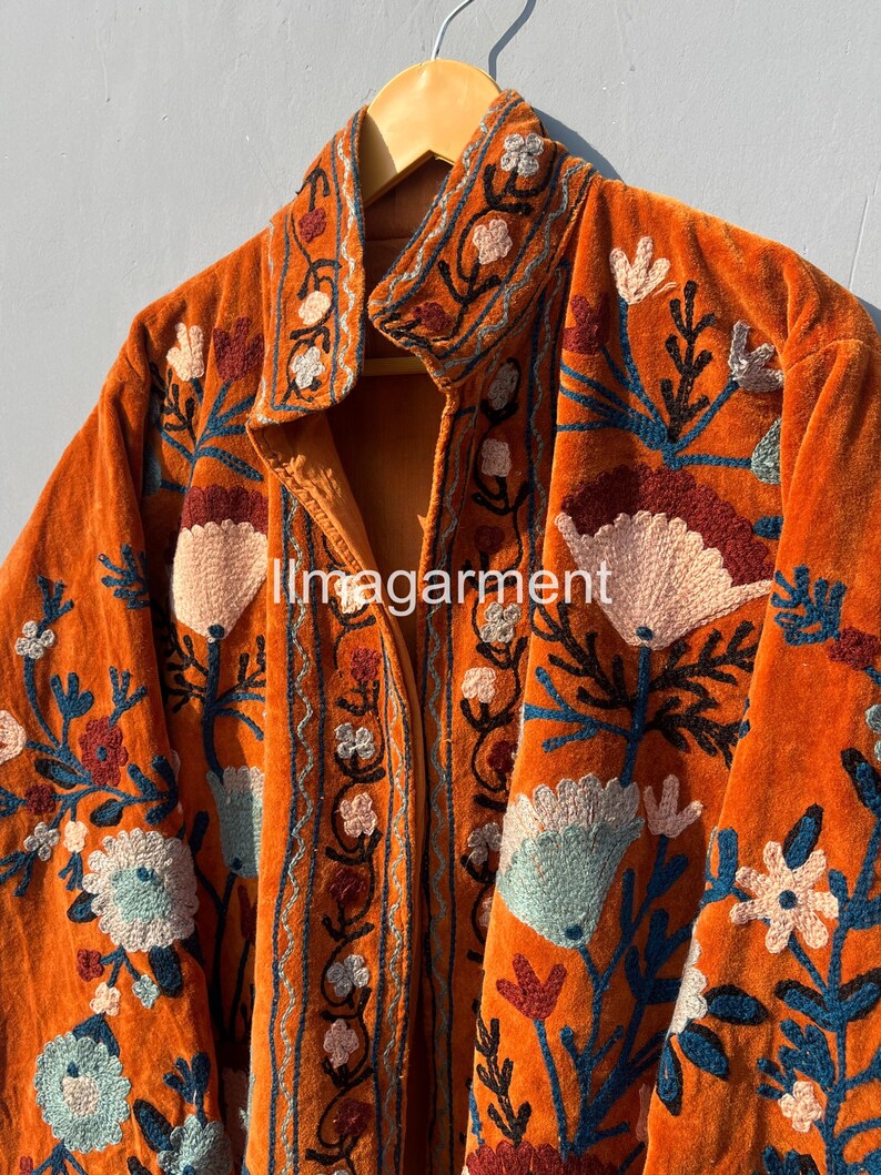 May include: A long, orange velvet jacket with intricate floral embroidery in shades of blue, pink, and brown. The jacket has a stand-up collar and a button closure.