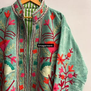 May include: A light teal jacket with floral embroidery in shades of pink, red, and orange. The jacket has a high collar and a striped green and white lining. The text "ilmagarment" is visible on the image.