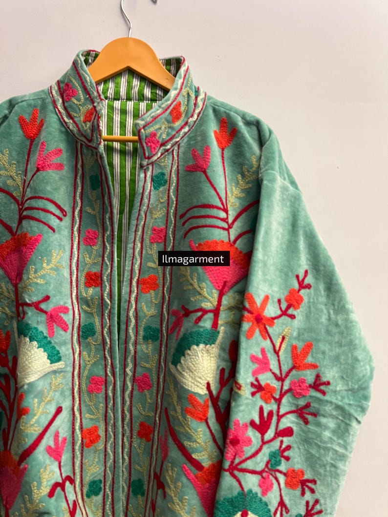 May include: A light teal jacket with floral embroidery in shades of pink, red, and orange. The jacket has a high collar and a striped green and white lining. The text "ilmagarment" is visible on the image.