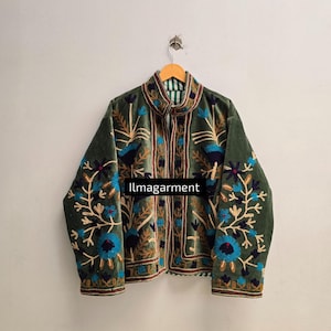 Puede incluir: Chaqueta de terciopelo verde con intrincados bordados florales en azul, marrón y dorado. La chaqueta tiene un cuello alto y un forro a rayas. El texto "llmagarment" es visible en la parte delantera.