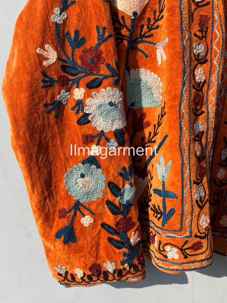 May include: Orange velvet jacket with intricate floral embroidery in shades of blue, pink, and green. The embroidery features a variety of flowers and leaves, creating a beautiful and intricate design.