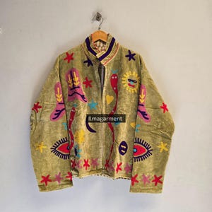 May include: An olive green jacket featuring colourful embroidered designs including a yellow sun, red snakes, blue stars, pink shapes, and eye motifs. The jacket has a high collar with purple trim and the text "ilmagarment" printed on the front.