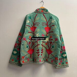 May include: A teal jacket with intricate floral embroidery in shades of red, pink, green, and white. The jacket is hanging on a wooden hanger against a plain white wall. The text "ilmagarment" is visible.
