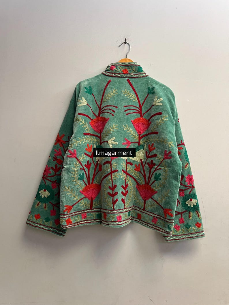 May include: A teal jacket with intricate floral embroidery in shades of red, pink, green, and white. The jacket is hanging on a wooden hanger against a plain white wall. The text "ilmagarment" is visible.
