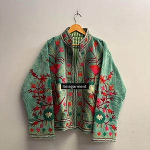 May include: A light green velvet jacket with intricate floral embroidery in shades of pink, red, and green. The jacket features a high collar and is lined with a striped fabric. The text "llmagarment" is visible on the front.