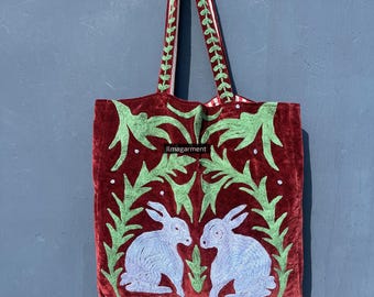 May include: A burgundy tote bag with green embroidered foliage and two light blue rabbits facing each other. The bag has long handles with a green leaf design and the text "ilmagarment".