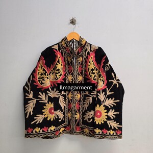 Könnte beinhalten: Schwarze Jacke mit aufwendiger Blumenstickerei in Rot, Gelb und Gold. Die Jacke hat einen hohen Kragen und wird auf einem Holzbügel präsentiert. Der Text "llmagarment" ist auf der Vorderseite der Jacke zu sehen.