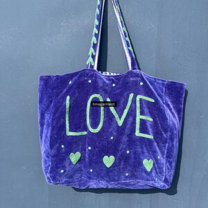 May include: A purple velvet tote bag with the word "LOVE" in green block letters. The bag has green heart and dot accents, and green and white patterned handles. The bag is hanging against a blue wall.