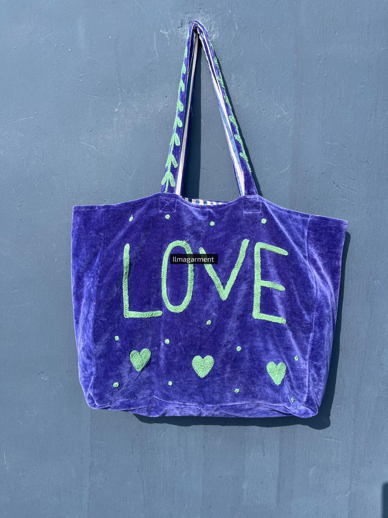 May include: A purple velvet tote bag with the word "LOVE" in green block letters. The bag has green heart and dot accents, and green and white patterned handles. The bag is hanging against a blue wall.