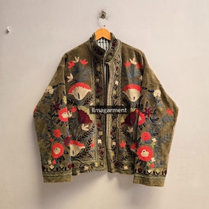 Può includere: Una giacca verde oliva con intricati ricami floreali nei toni del rosso, bianco e blu. Il capo presenta un colletto alto, maniche lunghe e un'apertura frontale. Il testo "Ilmagarment" è stampato in bianco al centro della giacca.