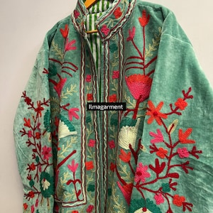 May include: A seafoam green jacket with intricate floral embroidery in shades of red, pink, and green. The jacket features a high collar and a striped lining. The text "ilmagarment" is visible on the front. The jacket is hanging on a wooden hanger.