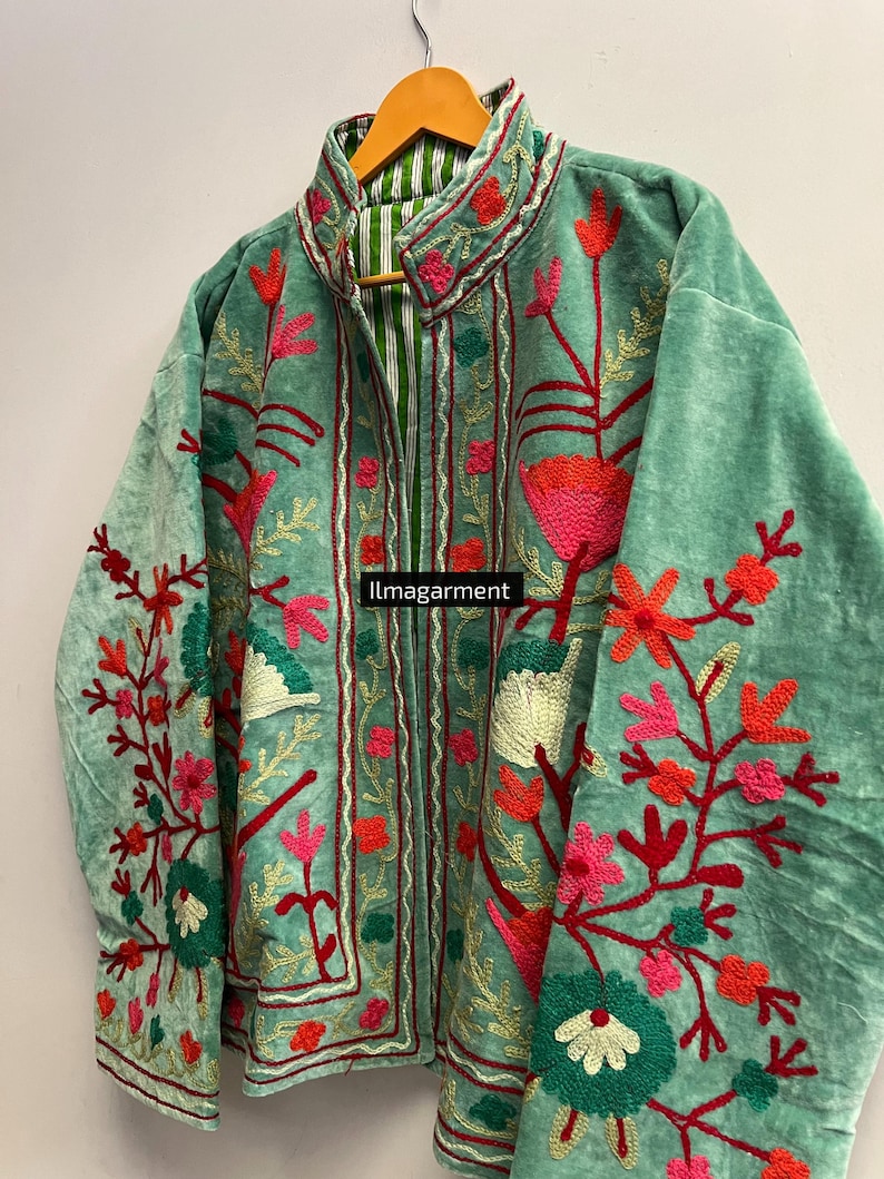 May include: A seafoam green jacket with intricate floral embroidery in shades of red, pink, and green. The jacket features a high collar and a striped lining. The text "ilmagarment" is visible on the front. The jacket is hanging on a wooden hanger.
