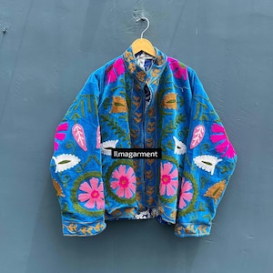 Pode incluir: Casaco azul com bordados florais em rosa, dourado e branco. O casaco tem gola alta e fecho de correr. O texto "llmagarment" é visível. Pendurado num cabide de madeira.