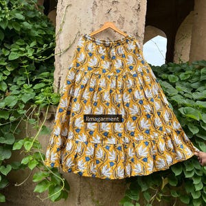 May include: Mustard yellow tiered skirt with a floral pattern in white and blue. The skirt is hanging on a wooden hanger. The text "llmagarment" is visible on the skirt.