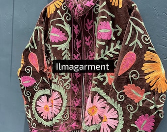 May include: A brown velvet jacket with intricate floral embroidery in pink, orange, and green. The jacket is hanging on a wooden hanger against a blue wall. The text "Ilmagarment" is visible on the jacket.