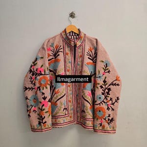 Puede incluir: Chaqueta rosa claro con intrincados bordados florales en tonos naranja, azul y rosa. La chaqueta tiene un cuello alto y una cremallera completa. El texto "llmagarment" es visible.