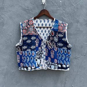 May include: A sleeveless vest with a patchwork design in shades of blue, red, and white. The vest features various patterns, including floral and elephant motifs. It has a tie closure and is hanging on a wooden hanger.