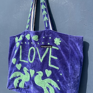 May include: A purple velvet tote bag with green embroidered details. The bag features the word "LOVE", two birds, and hearts. The handles are green with white stripes. The bag is a stylish accessory.