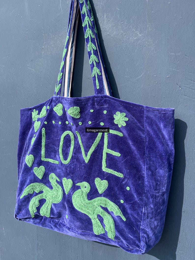 May include: A purple velvet tote bag with green embroidered details. The bag features the word "LOVE", two birds, and hearts. The handles are green with white stripes. The bag is a stylish accessory.
