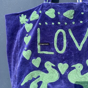 May include: A purple tote bag with green embroidered details. The bag features the word "LOVE" in large letters, along with hearts and bird designs. The bag has a soft, velvety texture and a sturdy handle.