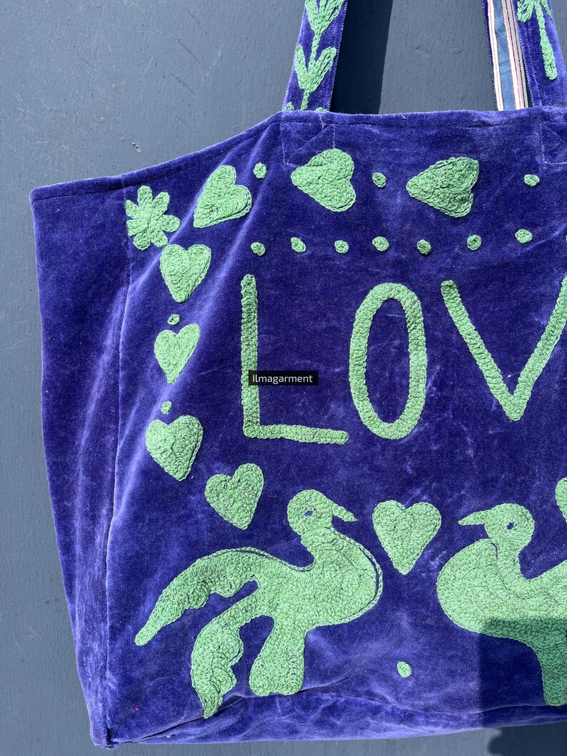 May include: A purple tote bag with green embroidered details. The bag features the word "LOVE" in large letters, along with hearts and bird designs. The bag has a soft, velvety texture and a sturdy handle.