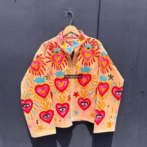 Könnte beinhalten: Hellpfirsichfarbene Jacke mit gestickten Herzen, Augen und Flammenmotiven. Die Jacke hat einen Kragen und lange Ärmel. Der Text "ilmagarment" ist auf der Jacke zu sehen.