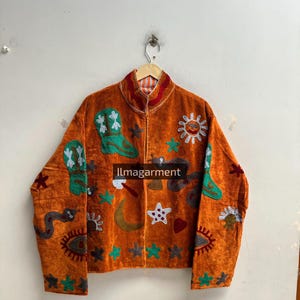 Op de afbeelding: Een oranje jas met geborduurde motieven, waaronder turquoise cowboylaarzen, een gele maansikkel, een witte zon, een grijze slang en diverse sterren. De tekst Ilmagarment staat in het midden. De jas heeft een hoge kraag.