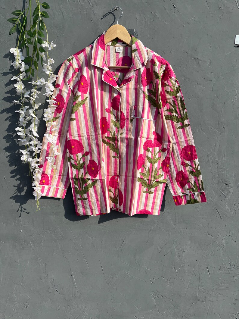 Puede incluir: Una camisa de manga larga a rayas rosa y blancas con estampado floral. La camisa tiene cuello y cierre de botones.