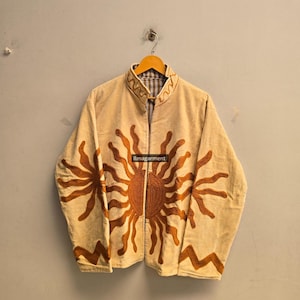 May include: A beige long-sleeve jacket featuring a prominent embroidered sun design in a dark orange hue across the front panels. The garment includes a mandarin collar with wavy line embroidery and the text ilmagarment printed on the center.