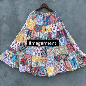 Könnte beinhalten: Ein mehrfarbiger Patchwork-Rock mit Stufen, der verschiedene Blumen- und geometrische Muster in Blau, Rosa, Gelb und Grün aufweist. Der Rock hängt an einem Holzbügel vor grauem Hintergrund. Der Text "llmagarment" ist sichtbar.