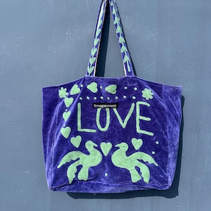 May include: A purple velvet tote bag with green embroidered details. The word "LOVE" is prominently displayed, along with heart and bird motifs. The handles are green with a decorative pattern.