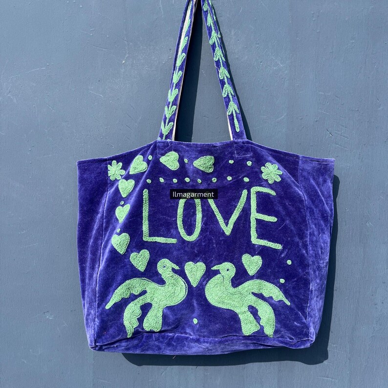 May include: A purple velvet tote bag with green embroidered details. The word "LOVE" is prominently displayed, along with heart and bird motifs. The handles are green with a decorative pattern.