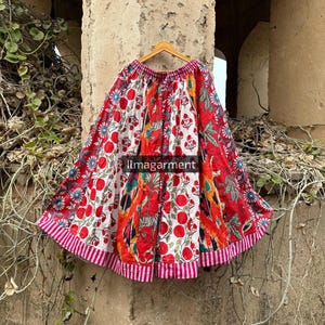 Peut inclure: Une jupe ample et colorée avec un motif patchwork. La jupe présente un mélange de motifs rouges, blancs et floraux, avec un ourlet rayé. Le texte "ilmagarment" est visible sur la jupe. La jupe est suspendue à un cintre en bois.