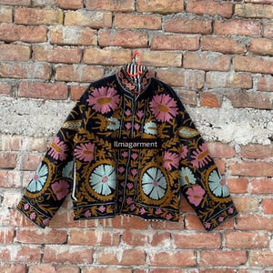 Peut inclure: Veste noire ornée de broderies florales colorées roses, bleues et dorées. La veste a un col montant et est suspendue à un cintre rayé rouge et blanc. Le texte "llmagarment" est visible.